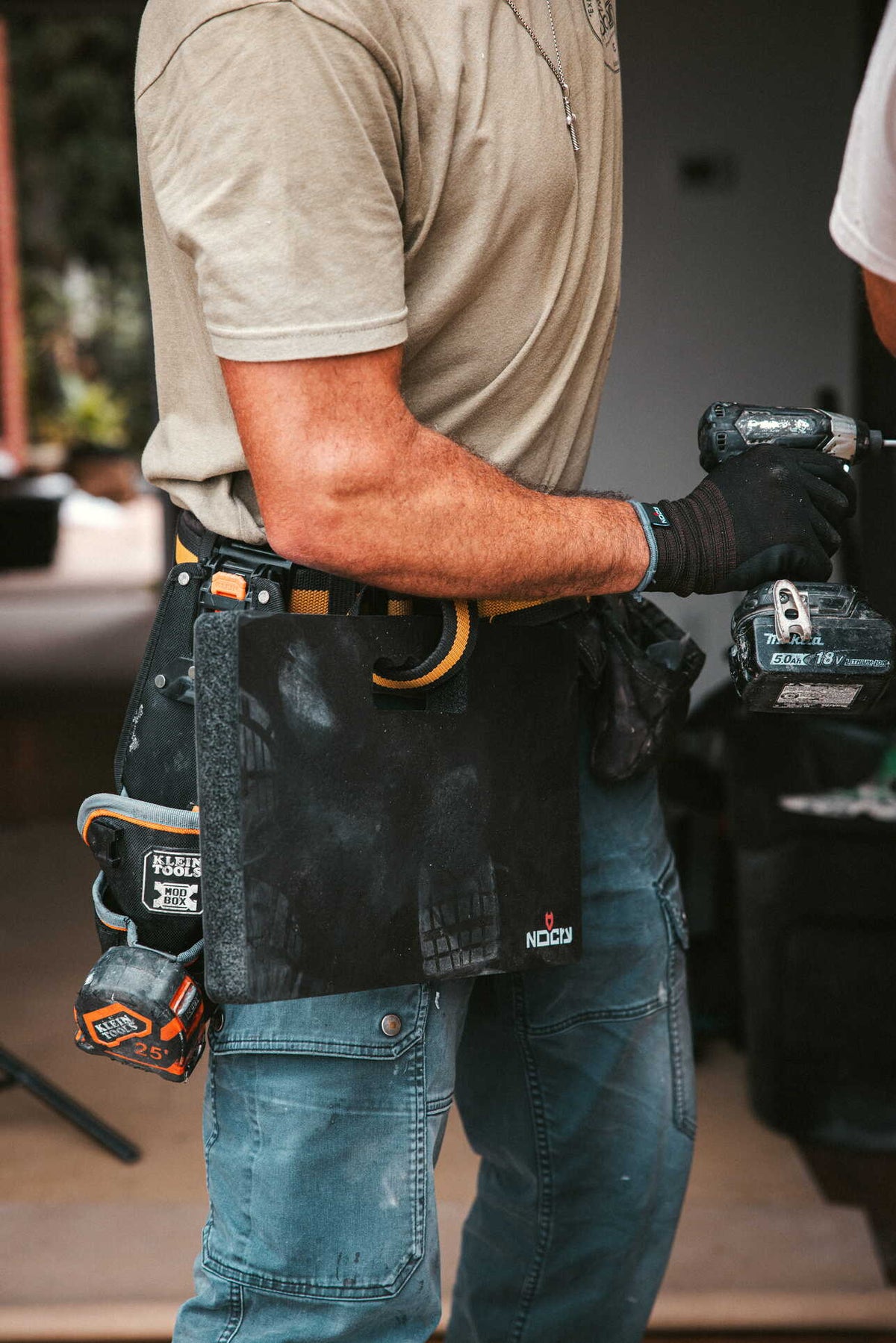 [First name], tile setter, adjusting the thigh strap on the leg, buckle and strap length visible, work pants, head in frame. Callout: "adjustable both sides".